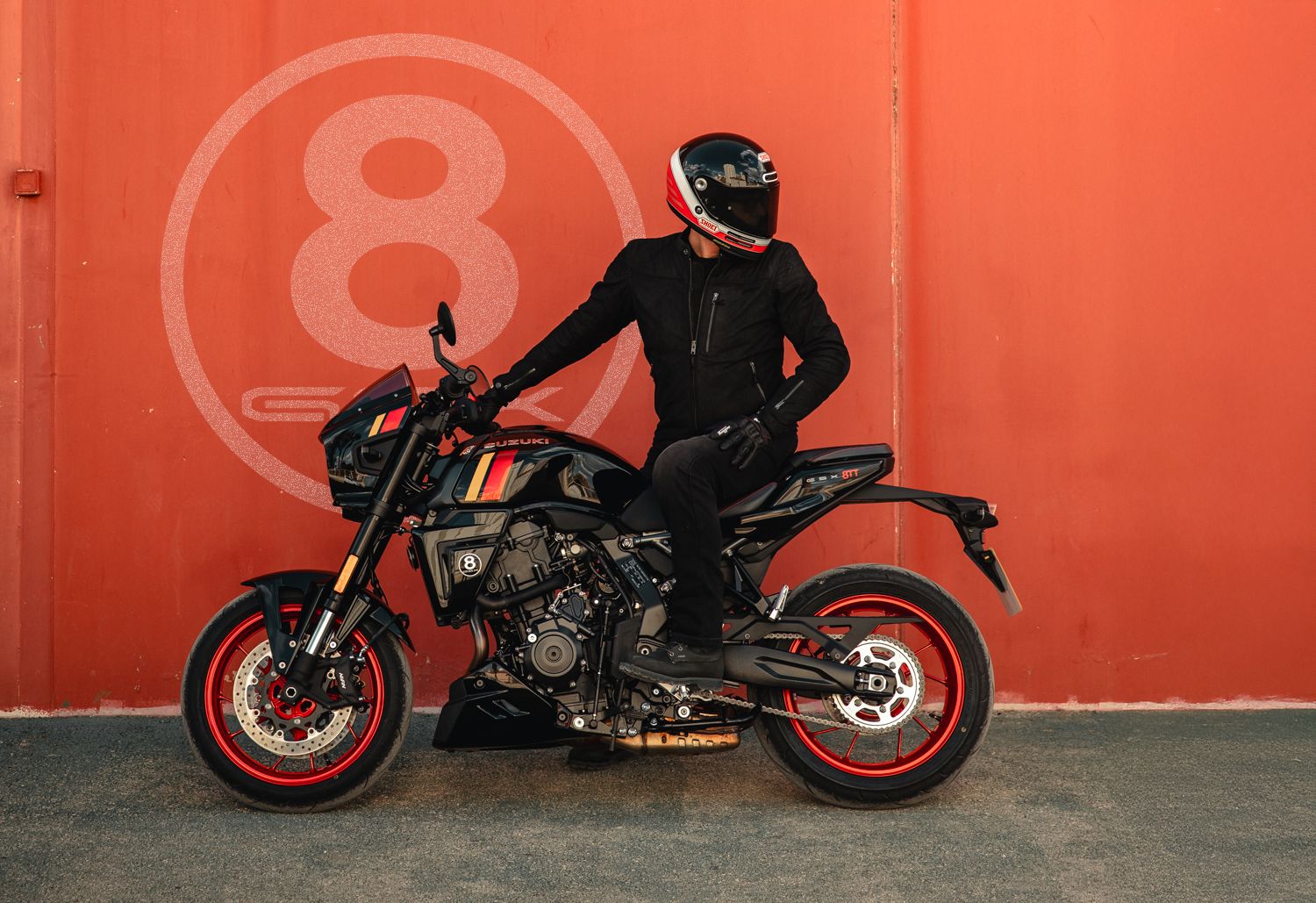 Fahrer in schwarzer Motorradkleidung sitzt auf einer Suzuki GSX-8TT im markanten Café-Racer-Design. Das Motorrad ist schwarz mit roten Felgen und sportlichen Farbakzenten auf dem Tank gestaltet. Im Hintergrund steht eine rote Wand mit großem 8-Ball-Logo.