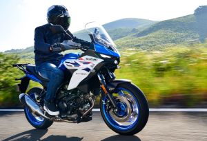Eine Person mit schwarzem Motorradhelm fährt auf einer blau-weißen Suzuki SV-7GX über eine Landstraße vor grünen Hügeln